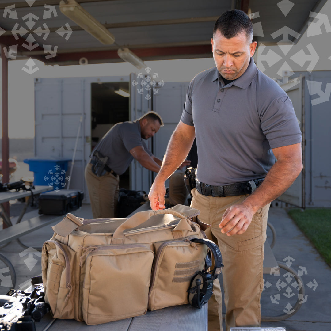 A man in a gray polo shirt and khaki cargo pants stands at a table with a tan tactical duffel bag in an outdoor storage or warehouse setting. Another man is visible in the background.