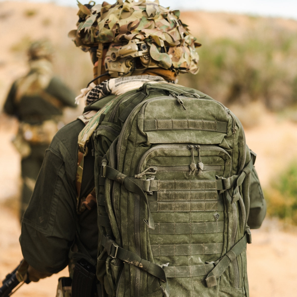 An olive green Specialist 1-Day Backpack shown from behind on a soldier in a desert setting, with another soldier blurred in the background.