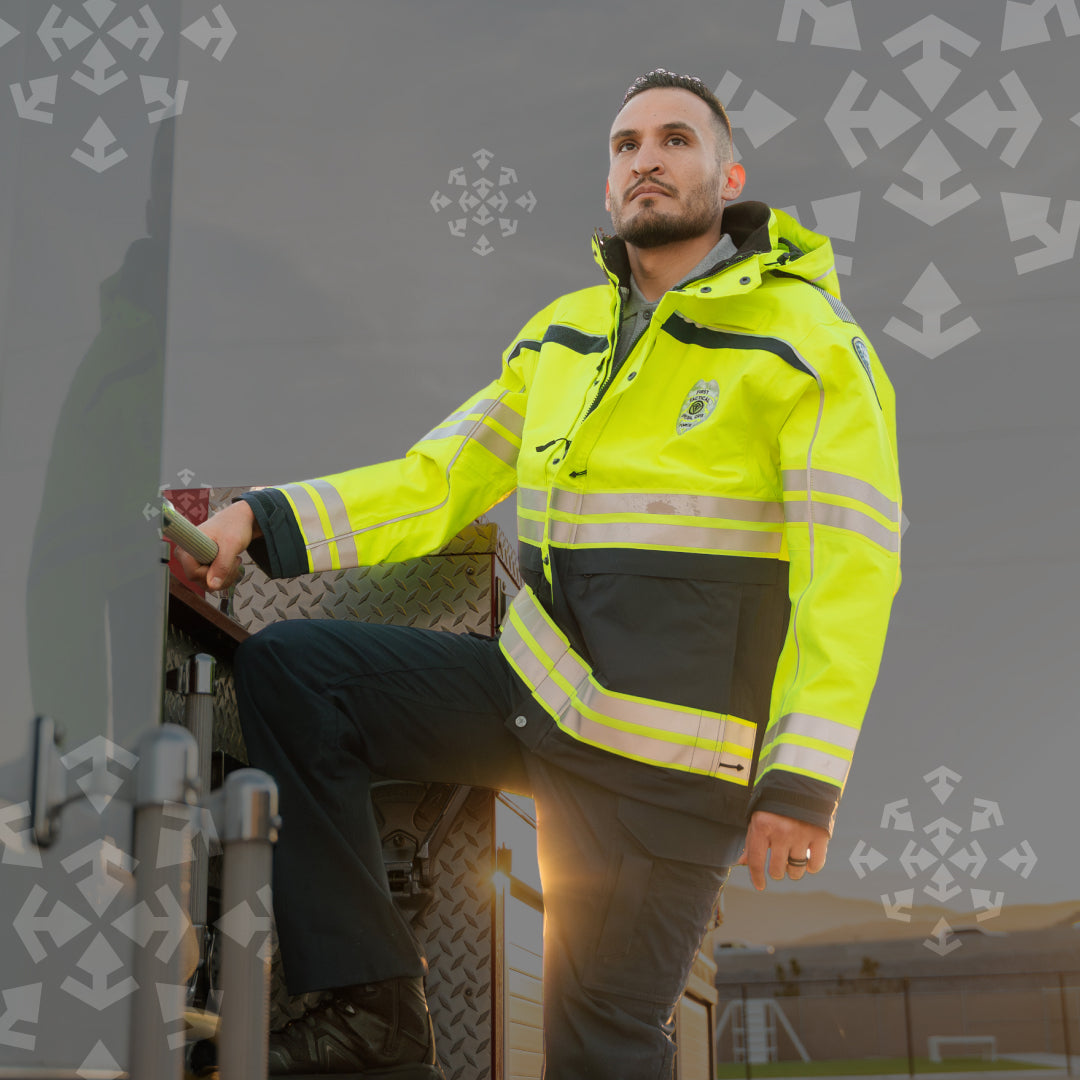A man wearing a high-visibility neon yellow safety jacket with reflective stripes and dark trousers, standing on the step of a truck in an outdoor setting.