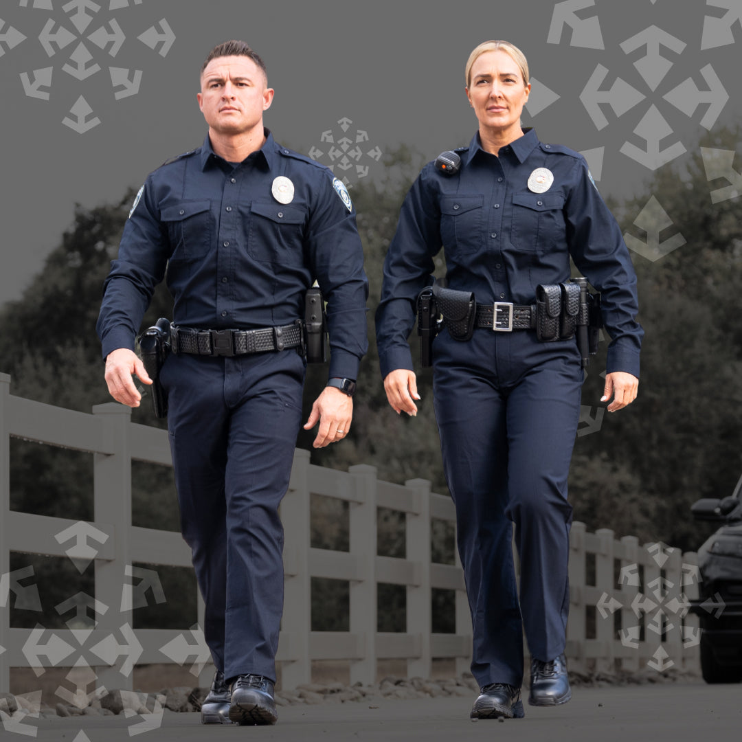 A man and a woman walking side by side in matching dark navy blue police uniforms with utility belts and badges, photographed outdoors on a road with a white fence and trees in the background.