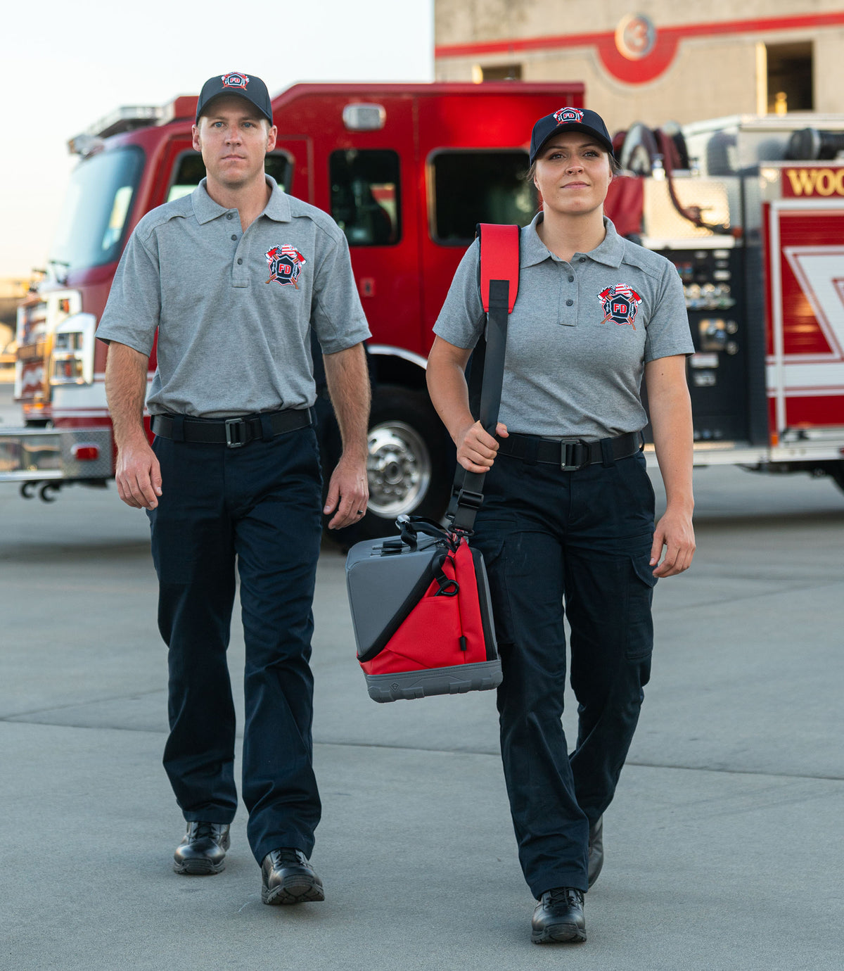 Two firefighters walking outdoors in matching gray polo shirts and dark pants, with a red fire truck in the background. One carries the red large jumper bag over her shoulder |color_black